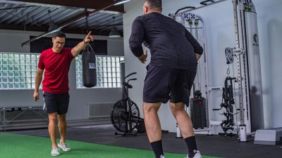 Back view of muscular man in sportswear doing exercises with trainer in modern fitness club